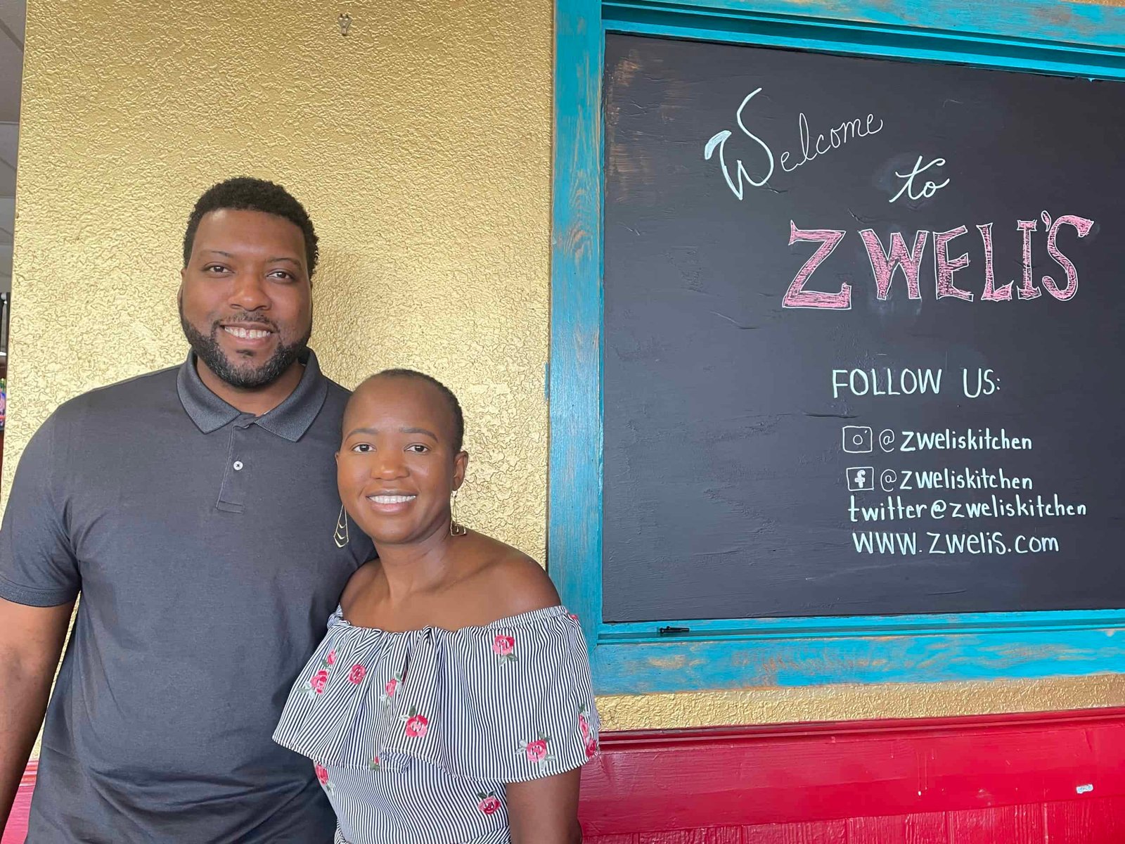 A couple standing next to a chalkboard in their restaurant that reads 