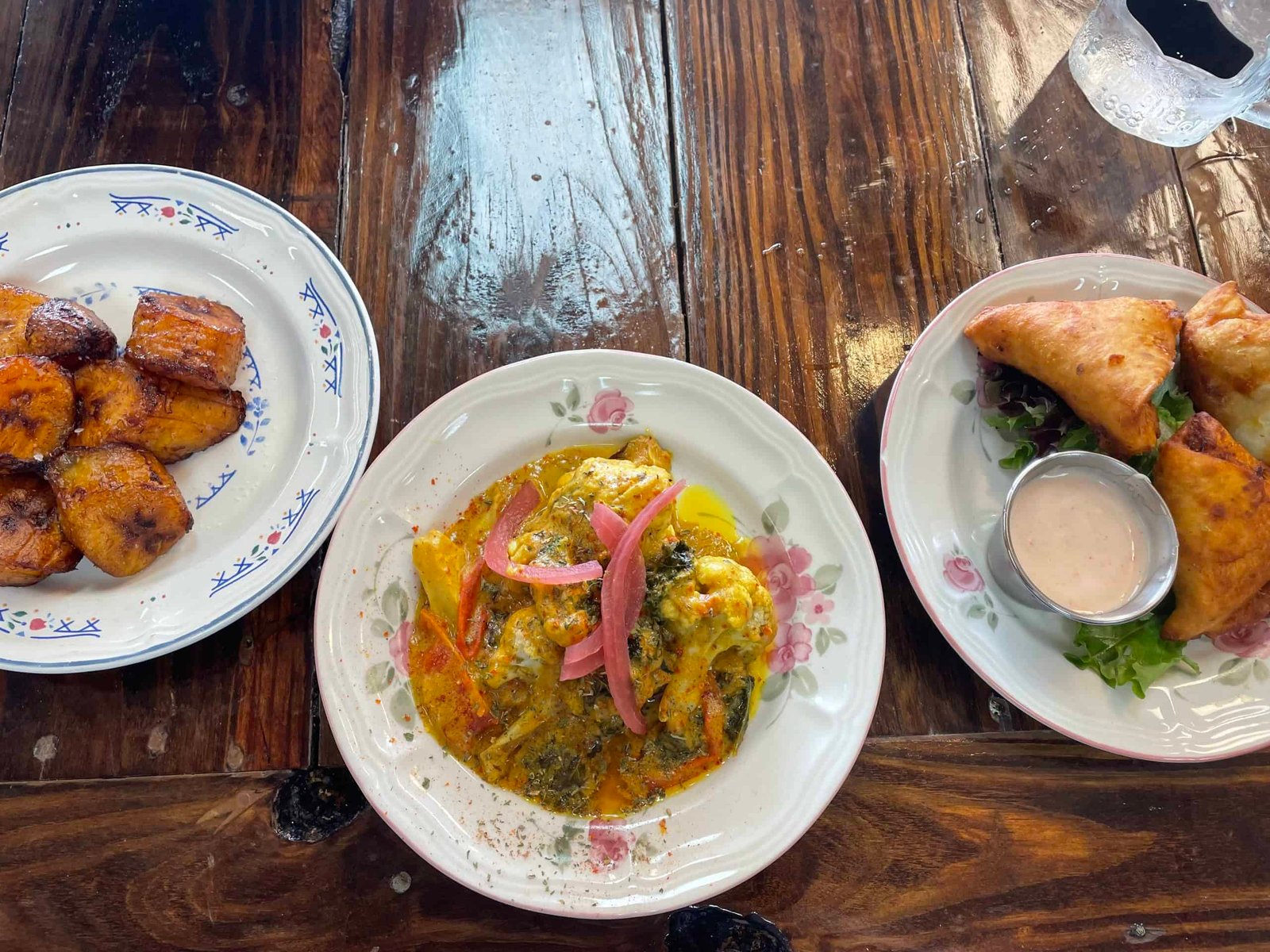 Image of three dishes including toasted plantains, colorful cauliflower stew with pickled onions, and samosas with sauce on the side.