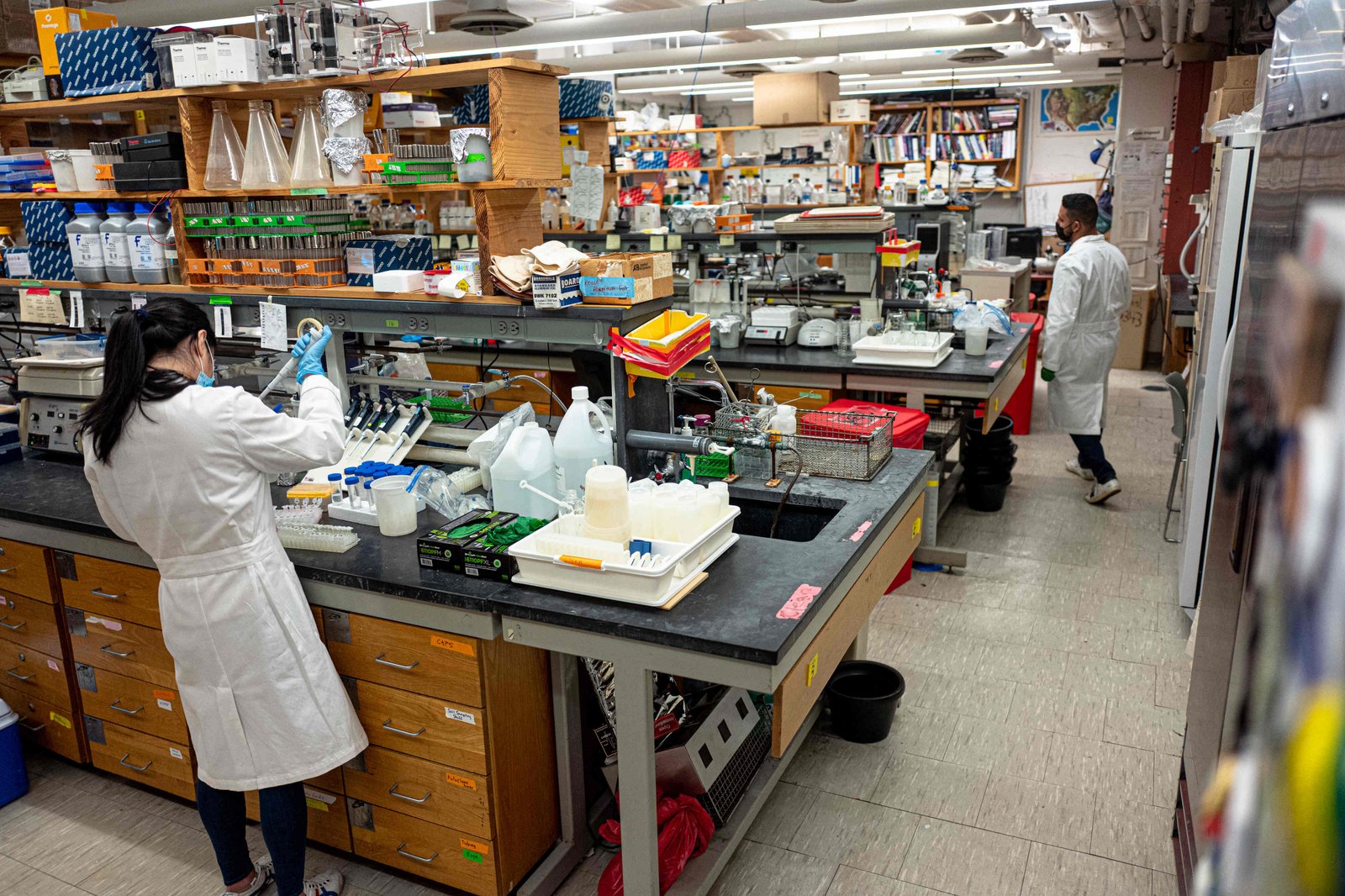 A wide, slightly elevated view of the Queens College lab space, whose shelves are replete with beakers, pipettes, weighing machines, notebooks and other assorted equipment and materials. At left, a lab technician in a white coat with her back to the viewer works at a lab bench.