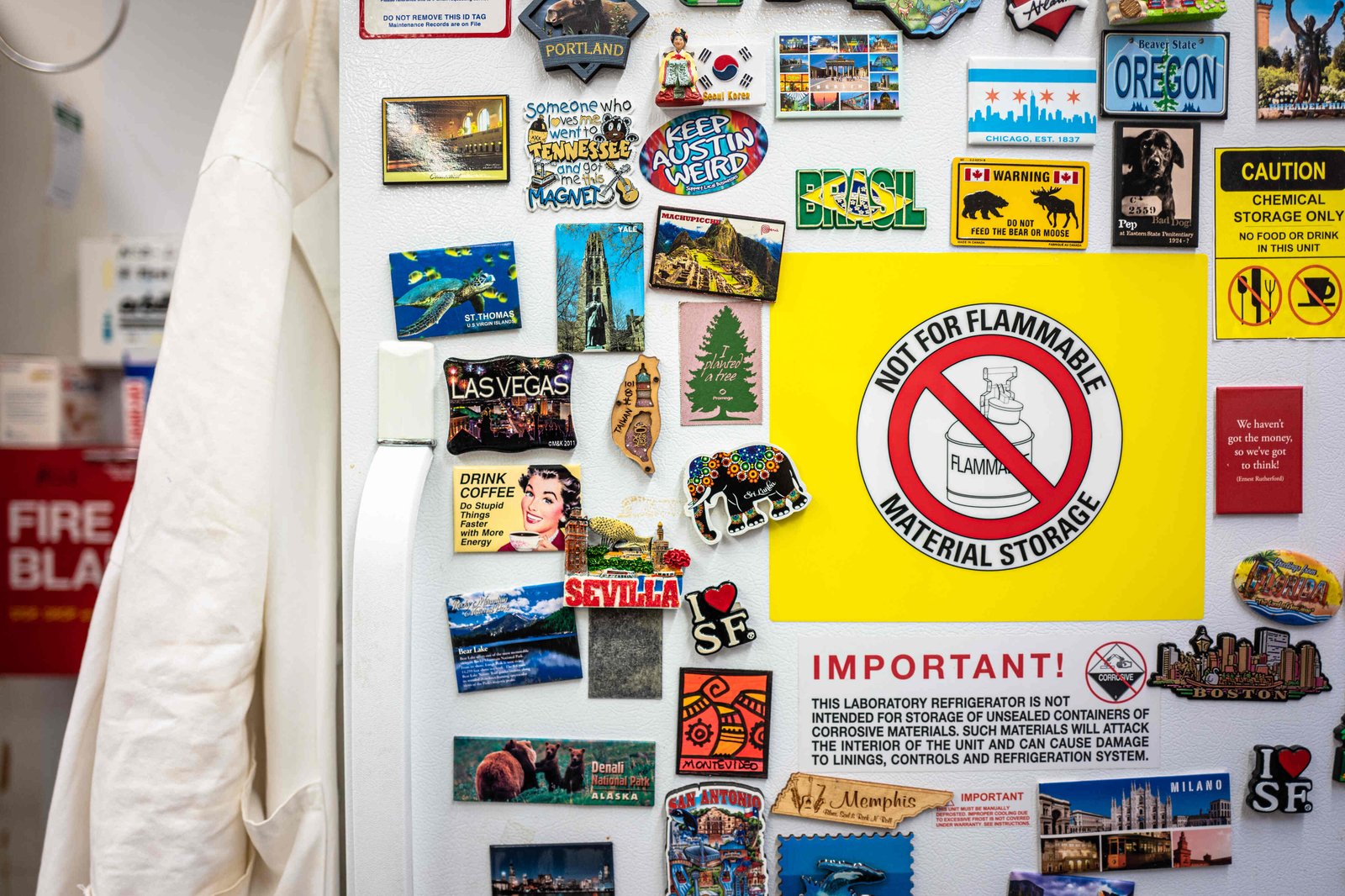 A close-up view of a white refrigerator door adorned with many magnets and signs. The most prominent signs read, “Not for flammable material storage,” and “Important! This laboratory refrigerator is not intended for storage of unsealed materials of corrosive materials.” The magnets are mostly colorful souvenir magnets from places like Las Vegas, Sri Lanka, Oregon, Machupiccu and Sevilla.