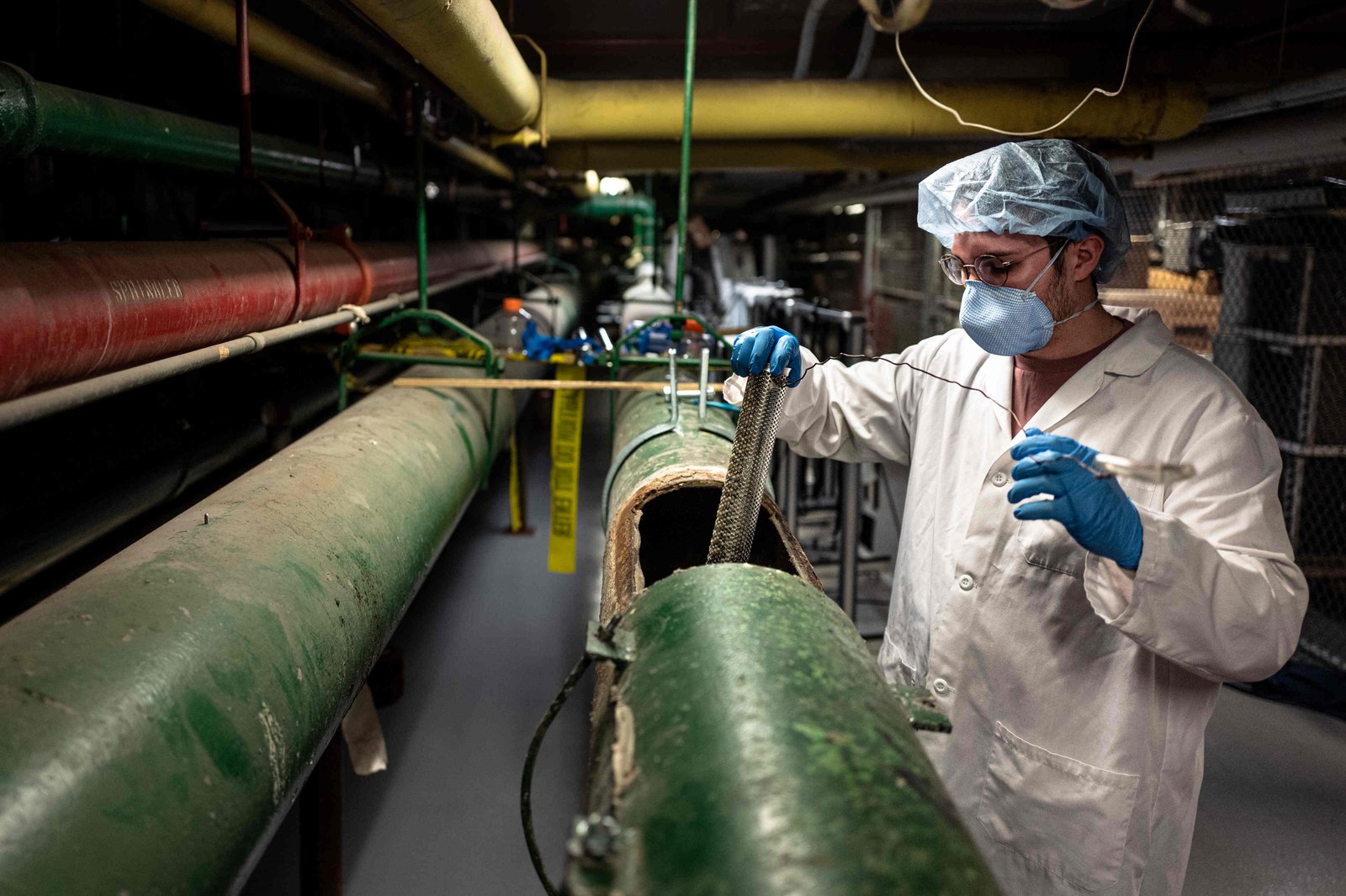 A lab technician in a hair net, white jacket and blue rubber gloves holds the Contraption, a porous metal cylinder with a wire attached to it, and guides it into the hole cut into the large green basement pipe.