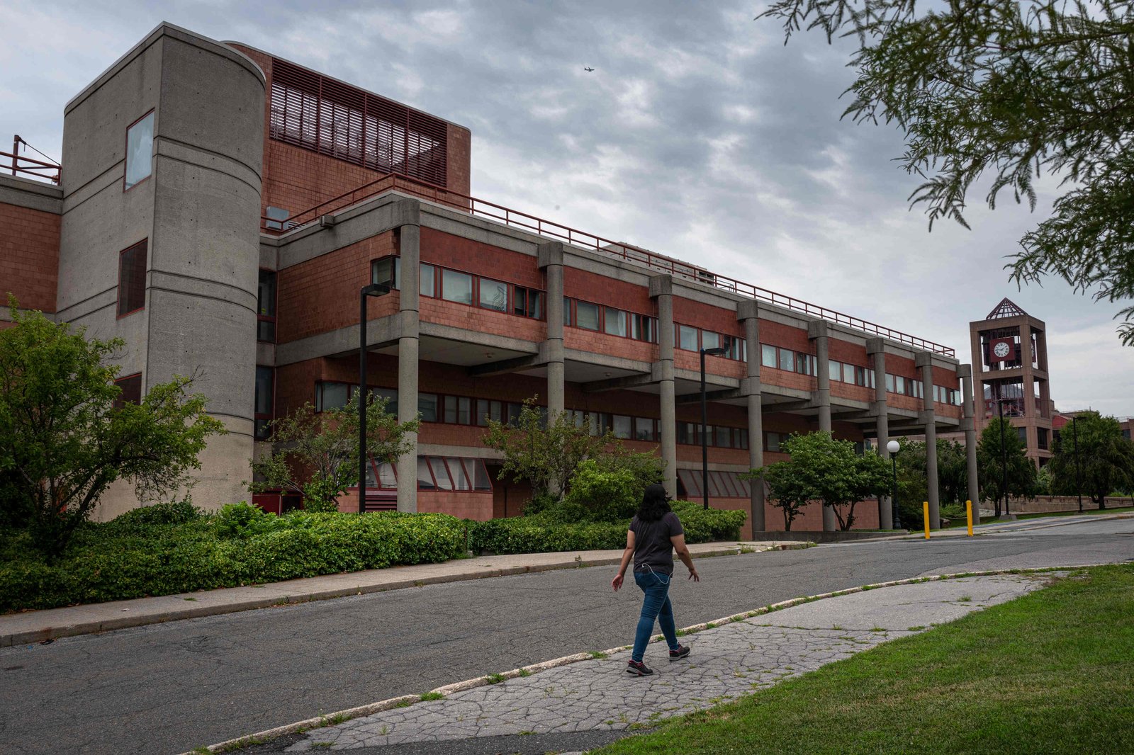 A street-level view of the Queens College building that contains the wastewater processing lab under an overcast sky. A person strolls by on the sidewalk.