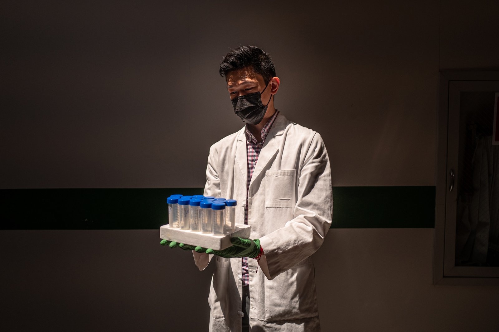 A researcher in a face mask and white lab coat and green rubber gloves holds a tray of wastewater samples that have blue tops.