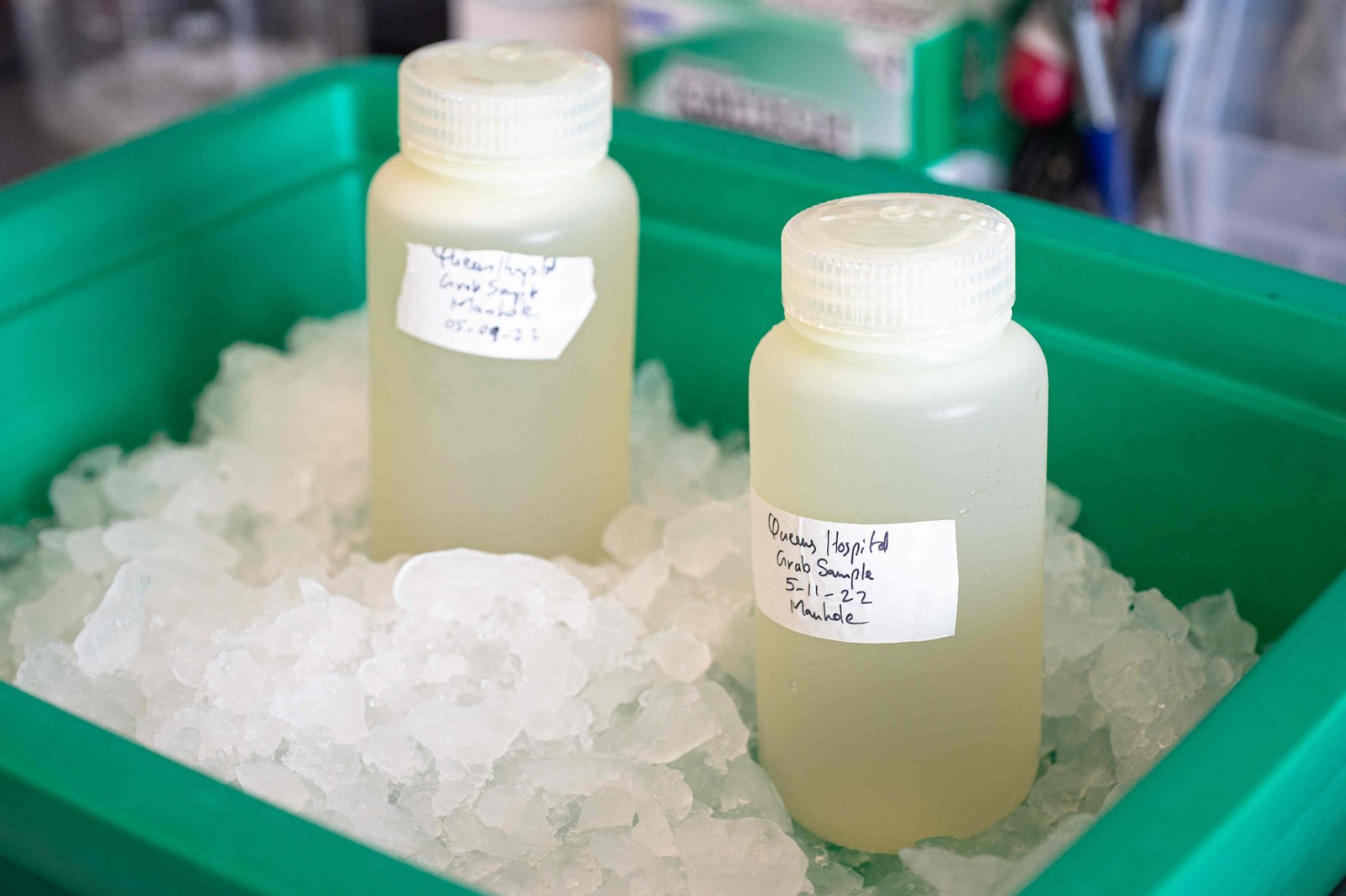 A close view of two plastic bottles containing sample water sit in a greenish tray in some ice. One of the bottles has a hand-written label reading “Queens Hospital Grab Sample, 5-11-22, Manhole.”