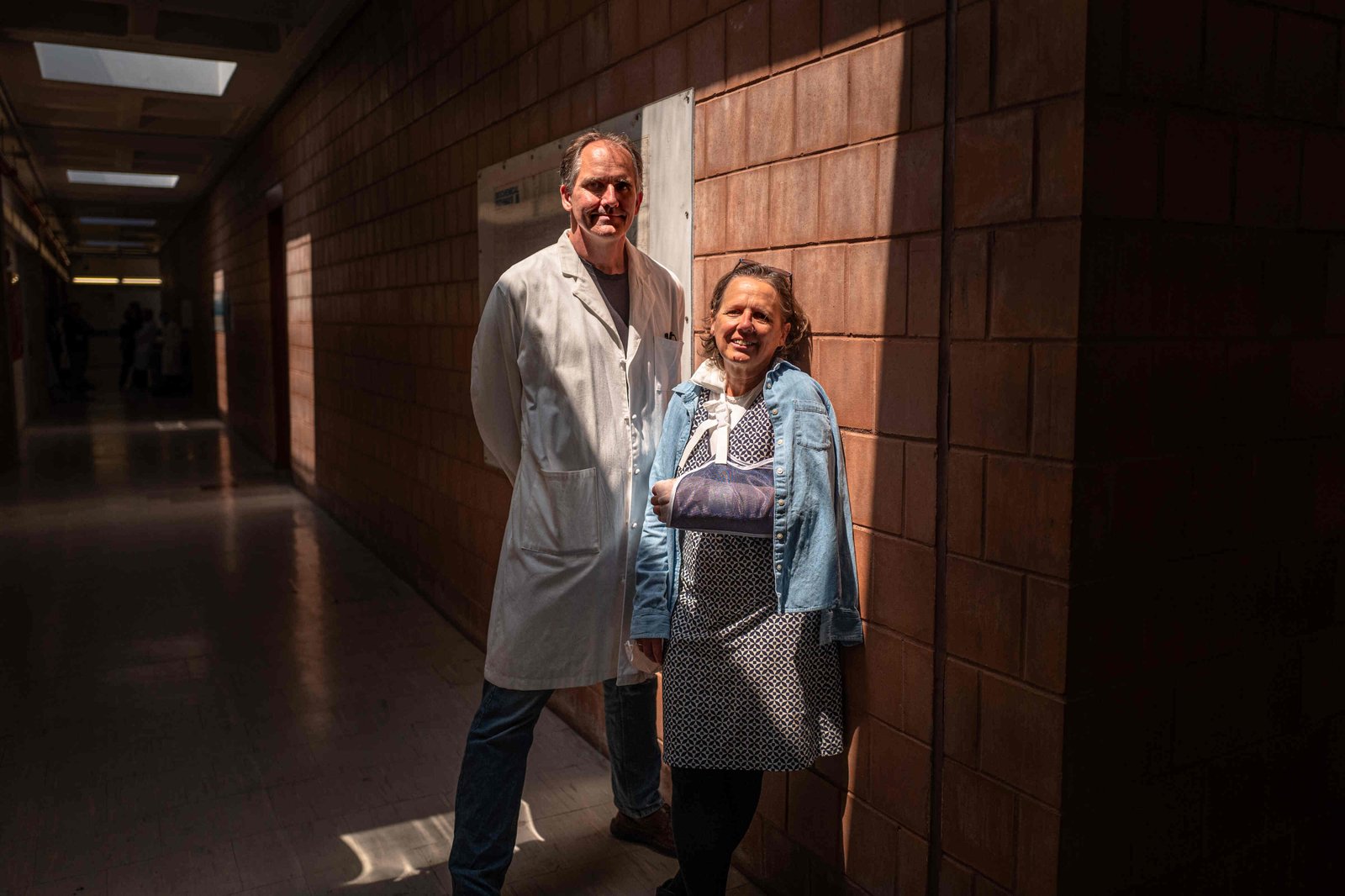 A double-portrait taken in a brick-walled hallway lit from above by skylights, showing John Dennehy, in a lab coat, left, and Monica Trujillo, who stand in a square of light cast from the skylight.