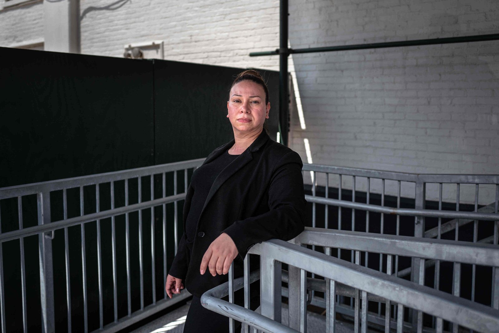 Leopolda Silvera, wearing a black suit jacket and a black shirt, leans against the railing of an accessibility ramp, looking directly at the viewer.