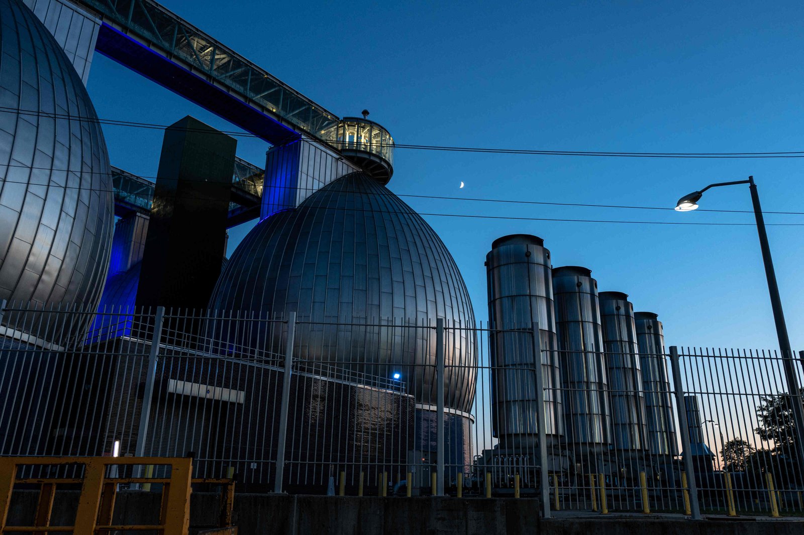 A ground-level view at dusk of the Newtown Creek Wastewater Treatment Plant, looking up at its large, bulbous and reflective “digester eggs” and a catwalk above them. There is a tall metal fence in the foreground, and a streetlight lit at right.