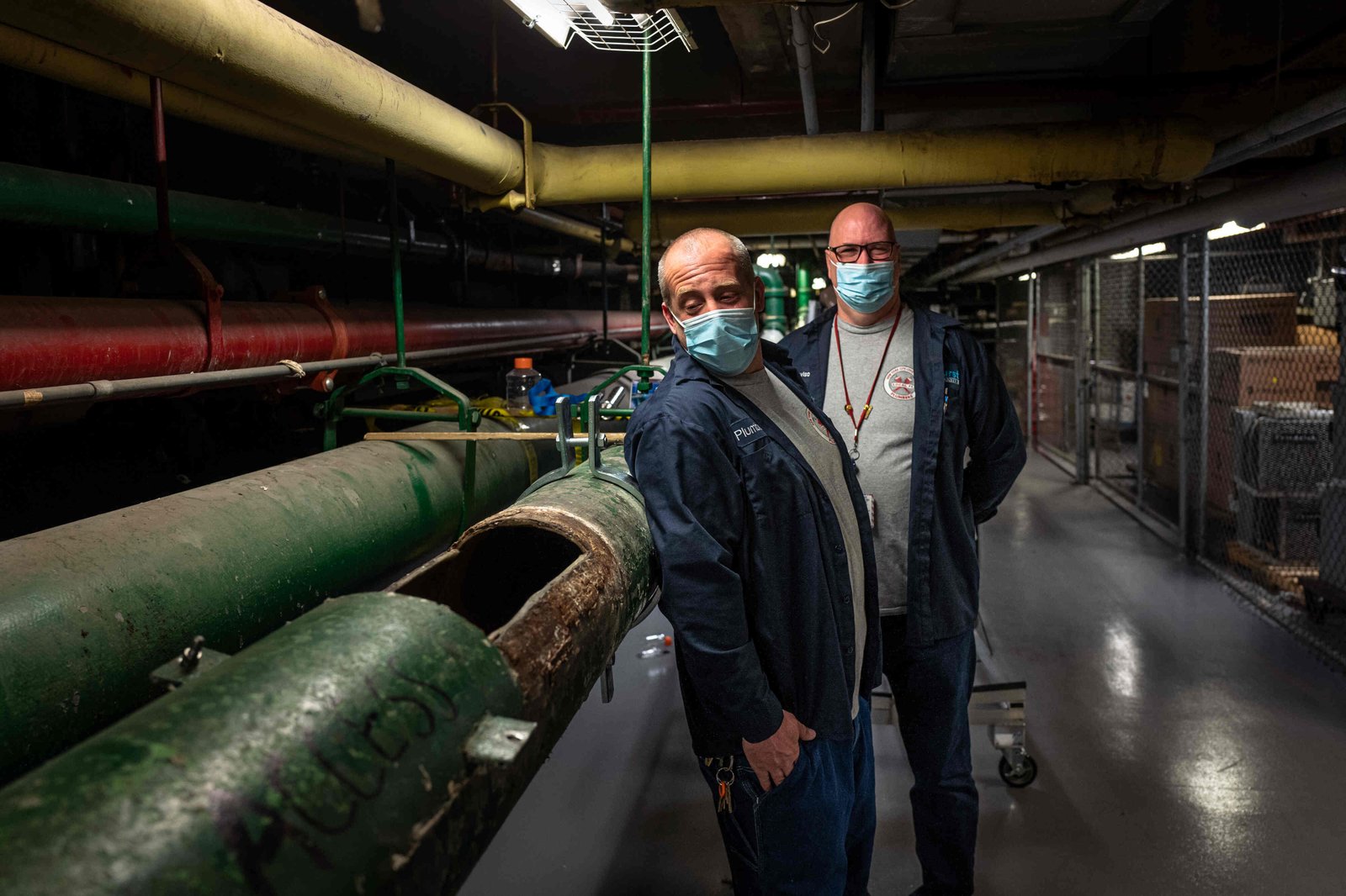 Two plumbers in blue jackets wearing face masks lean against one of two large green pipes in a basement. One pipe is partially cut open, and one of the plumbers peers in, while the other plumber looks at the camera.