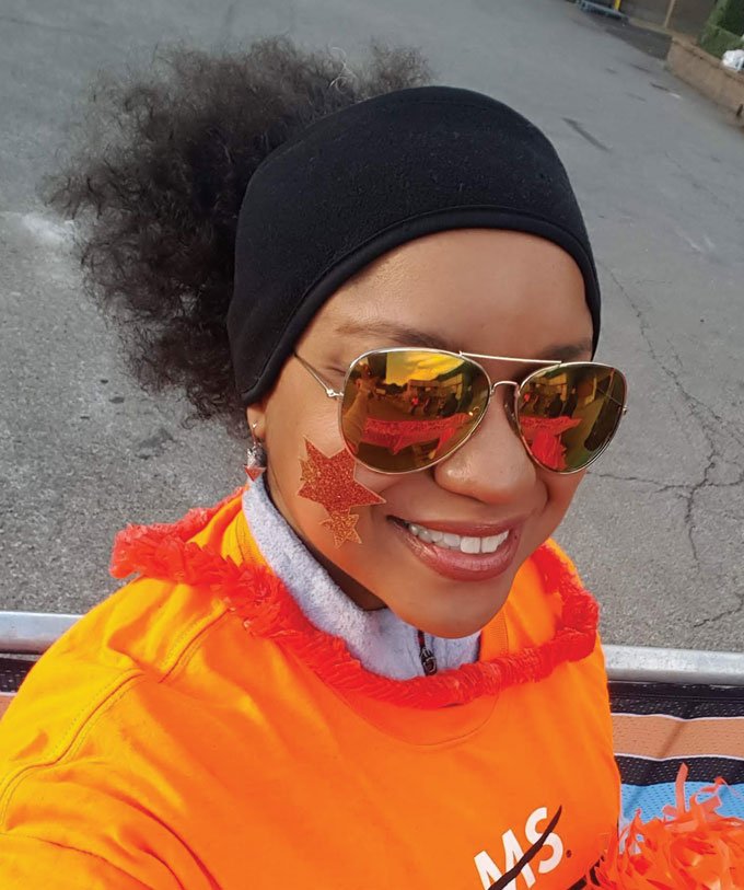 a photo of a smiling Black woman wearing sunglasses, a black headband, glitter stars on her cheek, and an orange shirt 