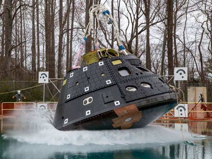 The Orion crew capsule, a cylindrical apparatus, being dropped into a water-filled basin with a forest in the background